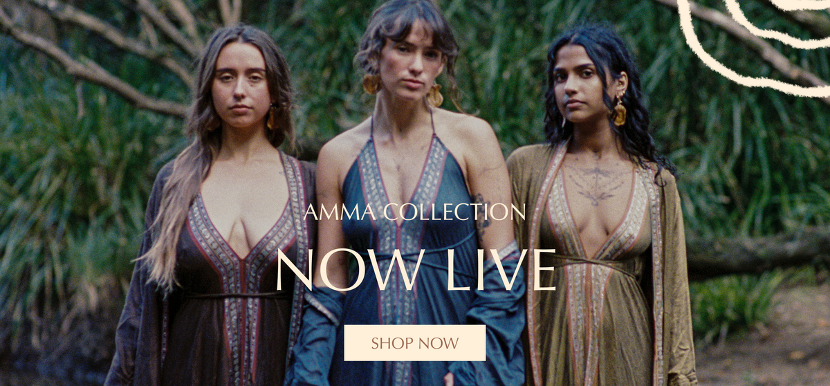 Three women standing outdoors in front of a forest background, wearing deep V-neck bohemian-style dresses in dark brown, blue, and beige. Text overlay reads 'AMMA COLLECTION NOW LIVE' with a 'SHOP NOW' button.